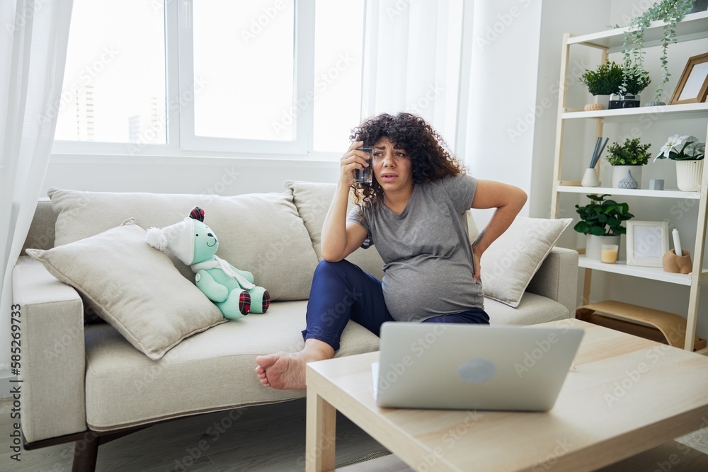 A pregnant woman sits on the couch with lower back pain and headache, strain on during pregnancy, drinks water and looks at the laptop. Lifestyle preparation for childbirth, last month of pregnancy A pregnant woman sits on the couch with lower back pain and headache, strain on during pregnancy, drinks water and looks at the laptop. Lifestyle preparation for childbirth, last month of pregnancy