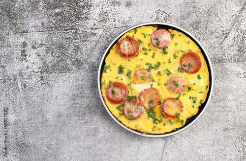 Omelet with herbs and sausage on a round plate on a dark grey background. Top view, flat lay