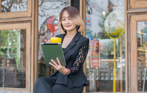 Beautiful businesswoman freelance sitting long wooden bench holding a coffee cup and using digital technology tablet VDO call conference with customer at the coffee shop outdoor while relaxing feeling