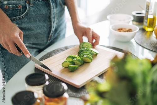 Young slim woman in white t-shirt and blue jeans cooking healthy food with cucumber in kitchen at home