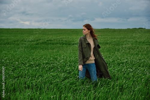a woman in a coat stands in the middle of a field in cloudy weather in a strong wind turned sideways to the camera