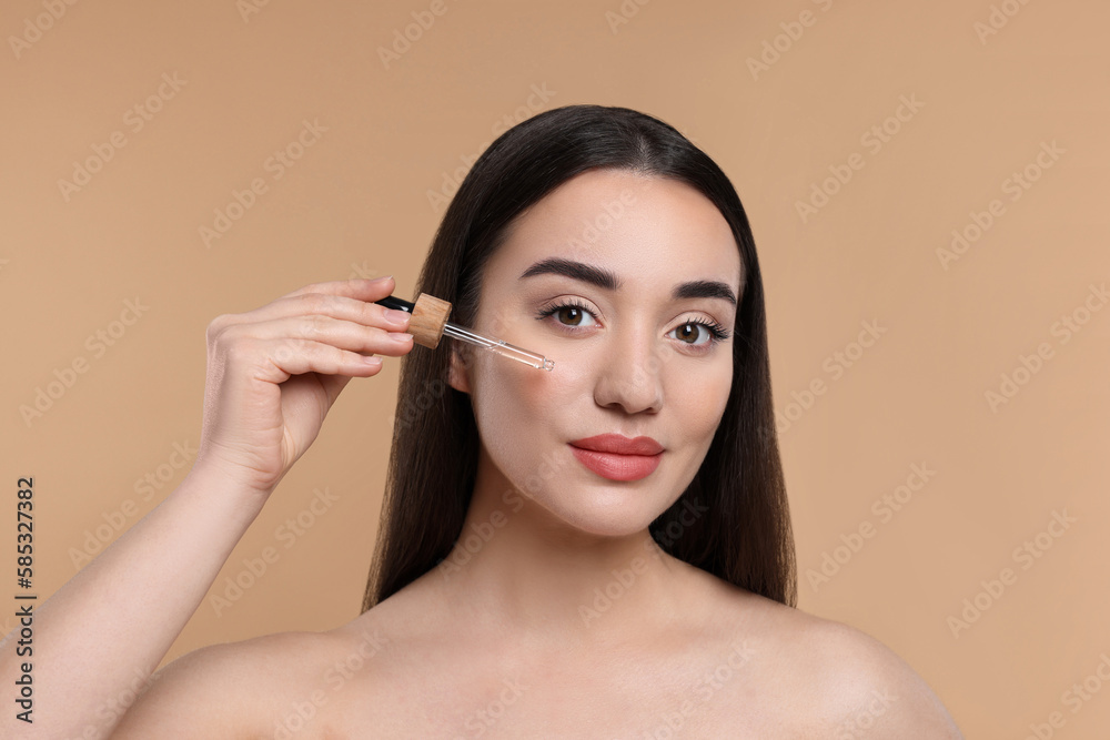 Young woman applying essential oil onto face on beige background