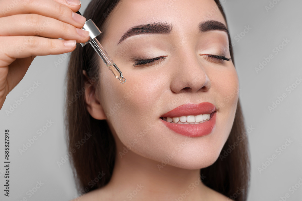 Happy young woman applying essential oil onto face on light grey background, closeup