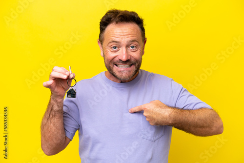 Middle age man holding home keys isolated on yellow background with surprise facial expression