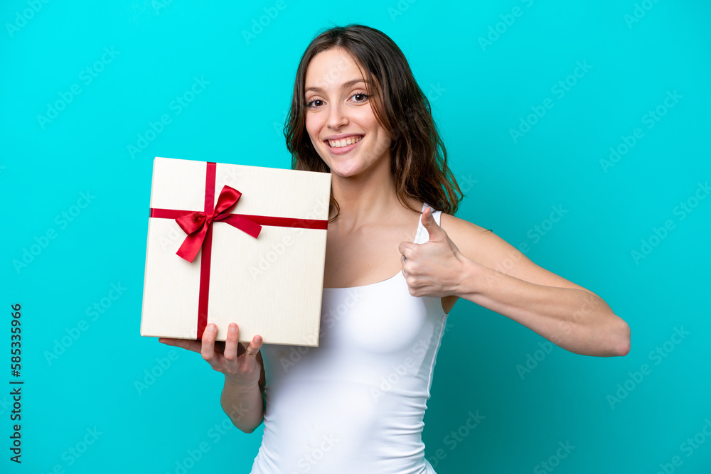 Young caucasian woman holding a gift isolated on blue background with thumbs up because something good has happened Young caucasian woman holding a gift isolated on blue background with thumbs up because something good has happened
