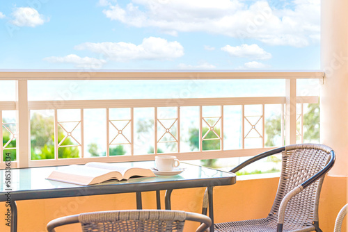 A cup of coffee and an open book over a balcony table. Background view of blue sky and sea. Summer vacation, time for relaxation, hobbies and recreation.