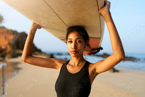 African american woman standing with surfboard on head on ocean beach. Black female surfer posing with surf board. Pretty multiethnic girl goes on surfing session on tropical location.