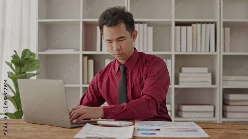 Businessman in red shirt working on laptop and paperwork that show cost planning accounting numbers tables on office space. Businessman in red shirt working on laptop and paperwork that show cost planning accounting numbers tables on office space.
