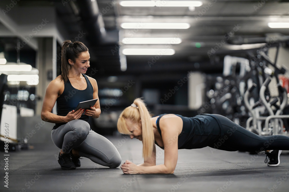 A happy female personal trainer is looking at the sportswoman who is doing  planks in a gym. Stock Photo
