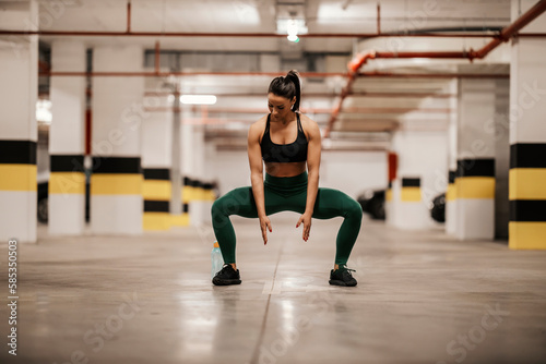 A muscular sportswoman in shape is doing squats in urban interior.