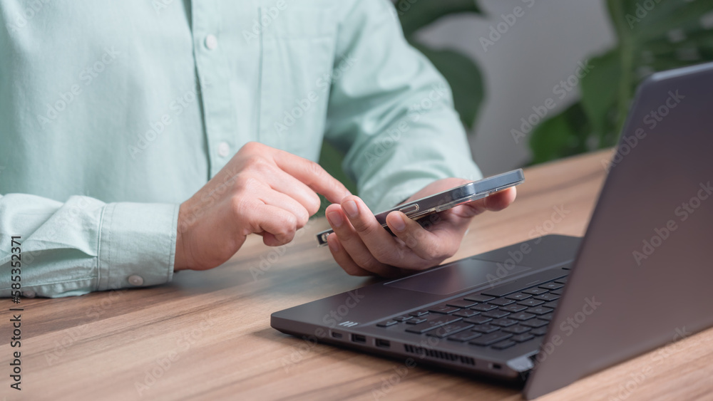 Male hand using a smartphone while working on computer laptop at office for investment analyzing company financial report balance with mobile phone. Concept of people are working with mobile devices. Male hand using a smartphone while working on computer laptop at office for investment analyzing company financial report balance with mobile phone. Concept of people are working with mobile devices.