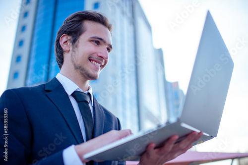 Happy confident businessman near a business center with a computer. Young smiling businessman in a suit is looking at a laptop. Cheerful businessman near a modern tall glass building with a computer.