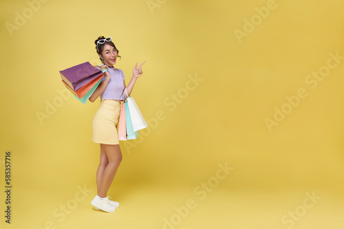 Cheerful happy thai asian woman enjoying shopping, she is carrying shopping bags pointing finger isolated on copy space yellow background.