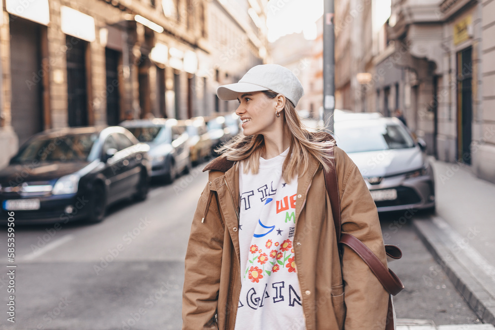 Happy girl with blonde hair in light cap and brown trench coat with handbag smiling outdoors, turn around and smiling. Stylish girl in fashion outfit walking over the city, look happy, hold bag. Happy girl with blonde hair in light cap and brown trench coat with handbag smiling outdoors, turn around and smiling. Stylish girl in fashion outfit walking over the city, look happy, hold bag.