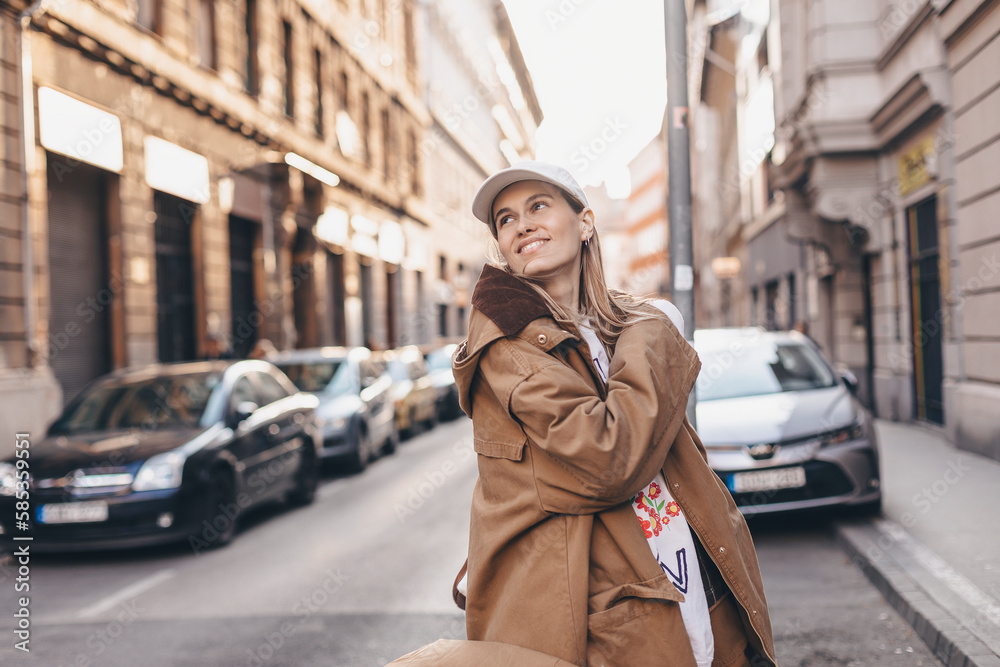 Happy girl with blonde hair in light cap and brown trench coat with handbag smiling outdoors, turn around and smiling. Stylish girl in fashion outfit walking over the city, look happy, hold bag.