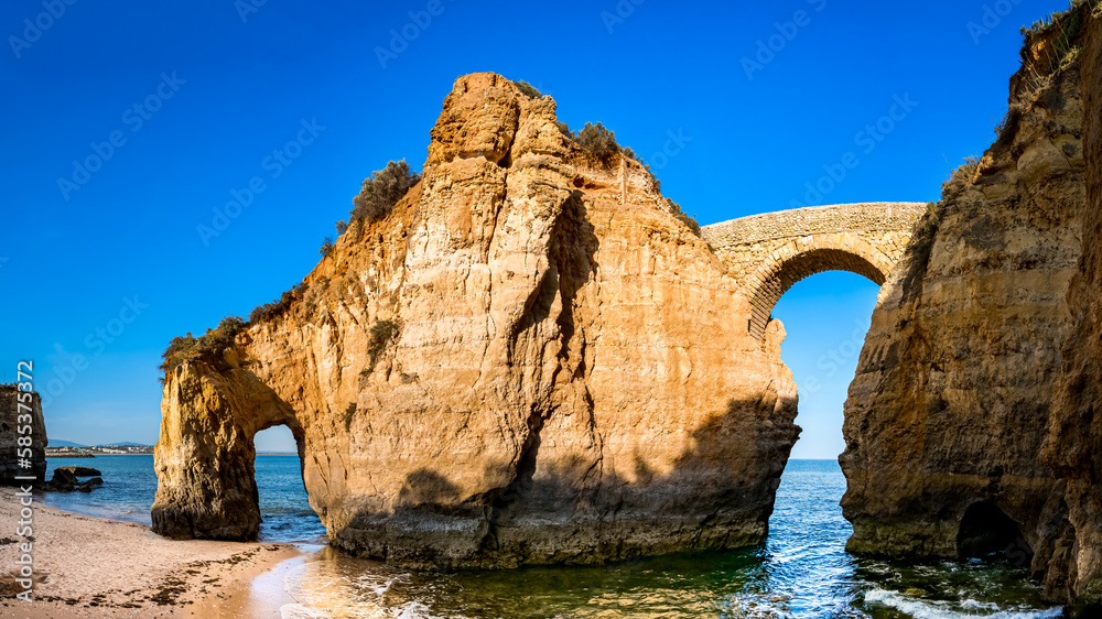 Historic Ponte Romana de Lagos stands gracefully amidst the golden hour glow at Praia dos Estudantes beach, connecting two cliffs and framing the horizon of the Atlantic Ocean in Algarve region.
