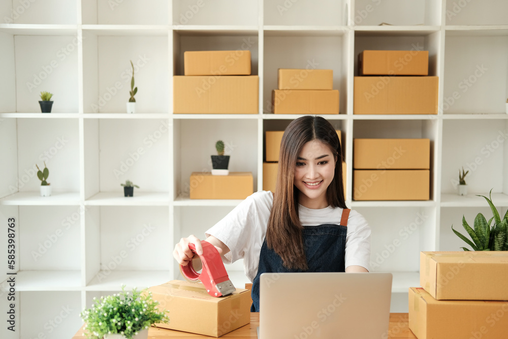 Startup small business, Young Asian woman owner using adhesive tape, packing carton parcel box working at the home office, seller prepares the delivery.