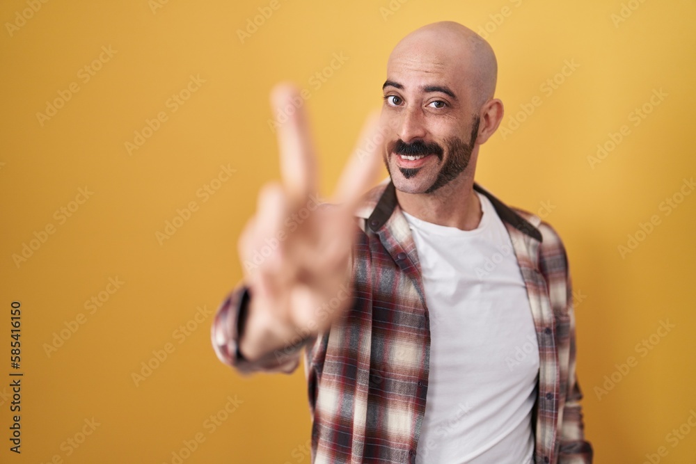 Fototapeta premium Hispanic man with beard standing over yellow background smiling looking to the camera showing fingers doing victory sign. number two.