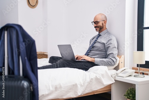 Young hispanic man business worker using laptop sitting on bed at hotel room