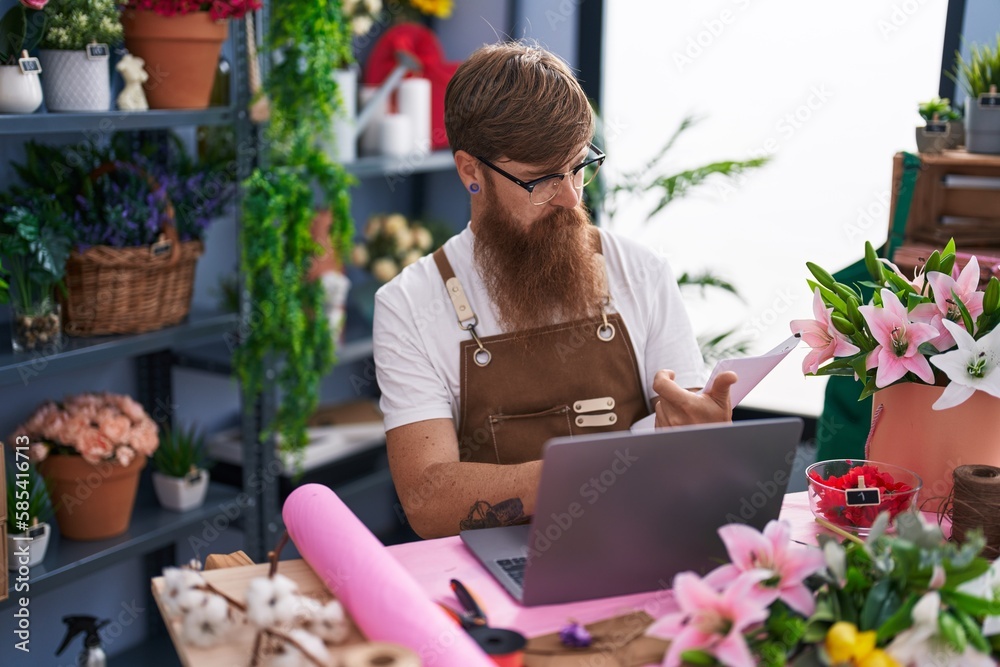 Young redhead man florist using laptop reading document at flower shop