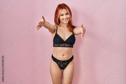 Young caucasian woman wearing lingerie over pink background looking at the camera smiling with open arms for hug. cheerful expression embracing happiness.