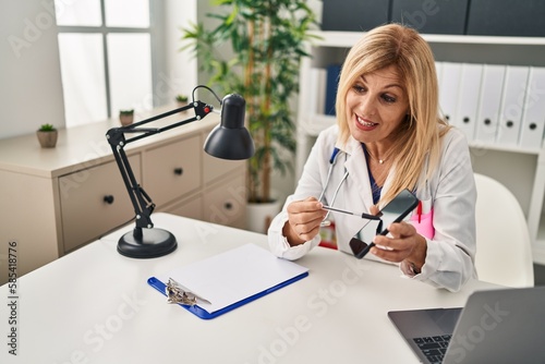 Middle age blonde woman wearing doctor uniform showing screen smartphone at clinic