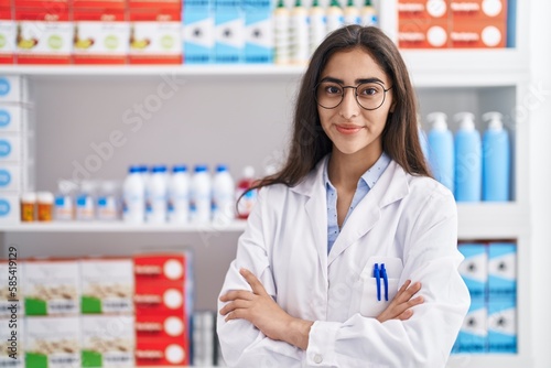 Young hispanic girl pharmacist standing with arms crossed gesture at pharmacy