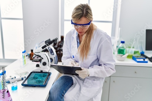 Young blonde woman wearing scientist uniform writing on clipboard looking embryion images at laboratory photo