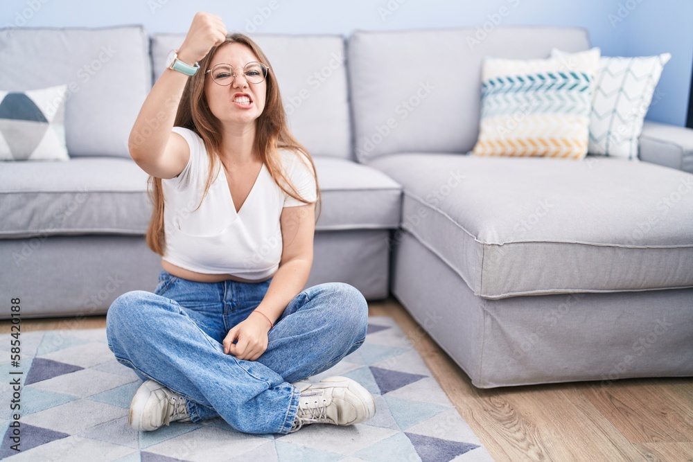 Young caucasian woman sitting on the floor at the living room angry and mad raising fist frustrated and furious while shouting with anger. rage and aggressive concept. Young caucasian woman sitting on the floor at the living room angry and mad raising fist frustrated and furious while shouting with anger. rage and aggressive concept.