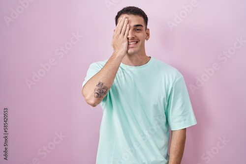 Handsome hispanic man standing over pink background covering one eye with hand, confident smile on face and surprise emotion.
