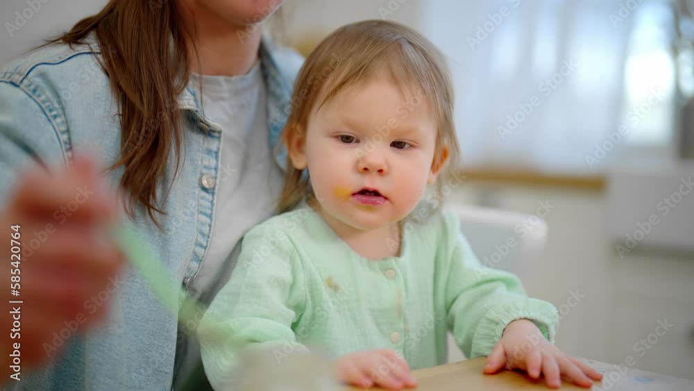 Mom feeds her baby playing with food. Toddler eats fruit or vegetable puree. Mother holds child on knees at table on kitchen. Raising child, kid's nutrition, motherhood, love care in family concept. Mom feeds her baby playing with food. Toddler eats fruit or vegetable puree. Mother holds child on knees at table on kitchen. Raising child, kid's nutrition, motherhood, love care in family concept.