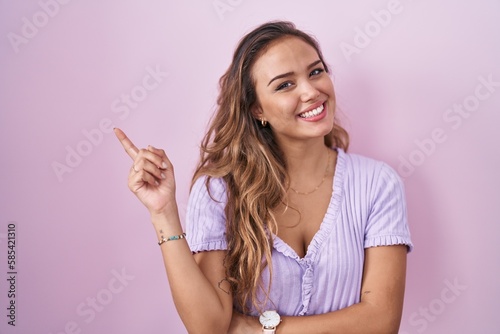 Young hispanic woman standing over pink background with a big smile on face, pointing with hand finger to the side looking at the camera. © Krakenimages.com