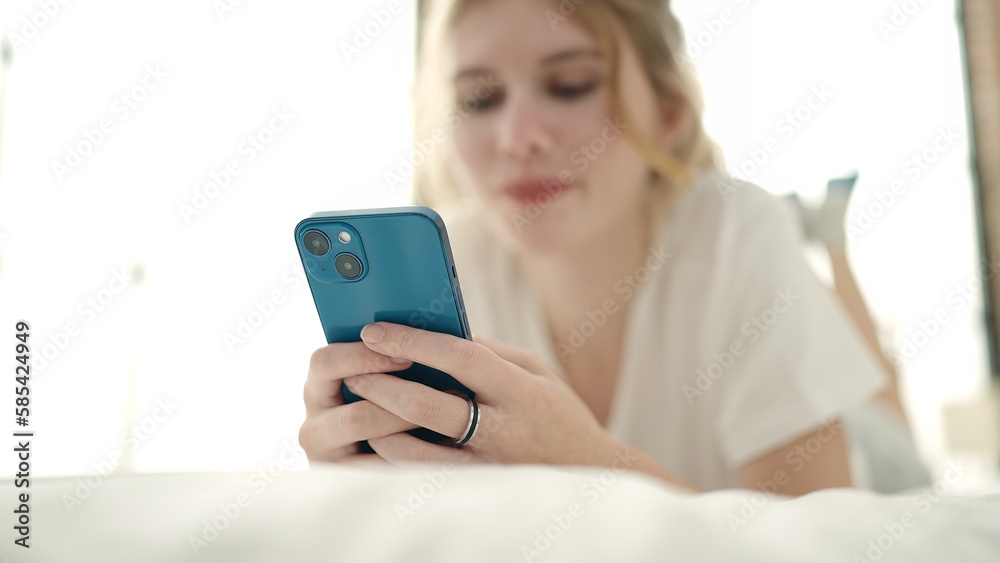 Young blonde woman using smartphone lying on bed at bedroom Young blonde woman using smartphone lying on bed at bedroom