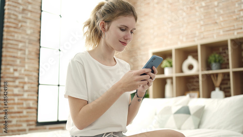 Young blonde woman using smartphone sitting on bed at bedroom