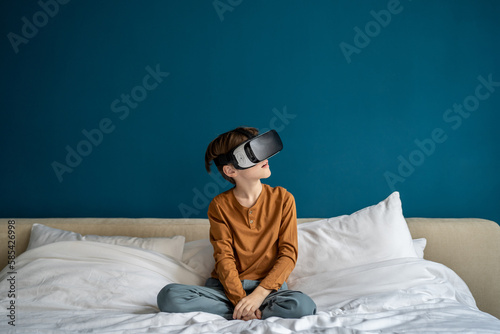 Interested kid boy using futuristic VR goggles interacting with virtual reality metaverse while sitting on bed at home. Schoolboy experiencing 3d headset immersive in game world in cyber space © DimaBerlin