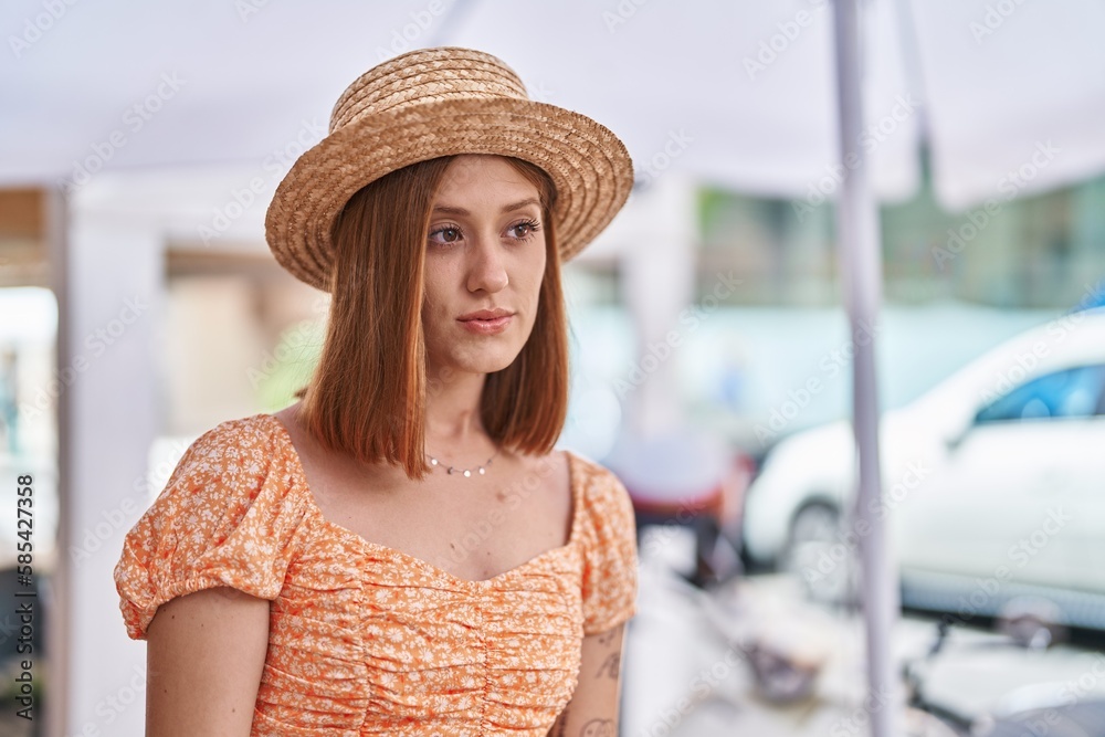 Young redhead woman tourist wearing summer hat standing with relaxed expression at street