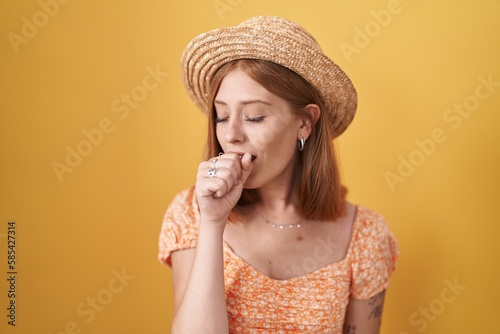 Young redhead woman standing over yellow background wearing summer hat feeling unwell and coughing as symptom for cold or bronchitis. health care concept.