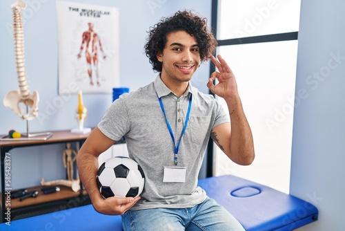 Hispanic man with curly hair working as football physiotherapist smiling positive doing ok sign with hand and fingers. successful expression.