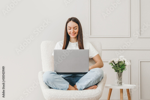 Caucasian student girl sits in a chair uses a laptop and looks at the monitor screen © Evgenia