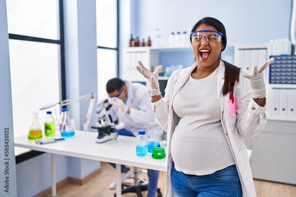 Young hispanic woman expecting a baby working at scientist laboratory celebrating mad and crazy for success with arms raised and closed eyes screaming excited. winner concept Young hispanic woman expecting a baby working at scientist laboratory celebrating mad and crazy for success with arms raised and closed eyes screaming excited. winner concept