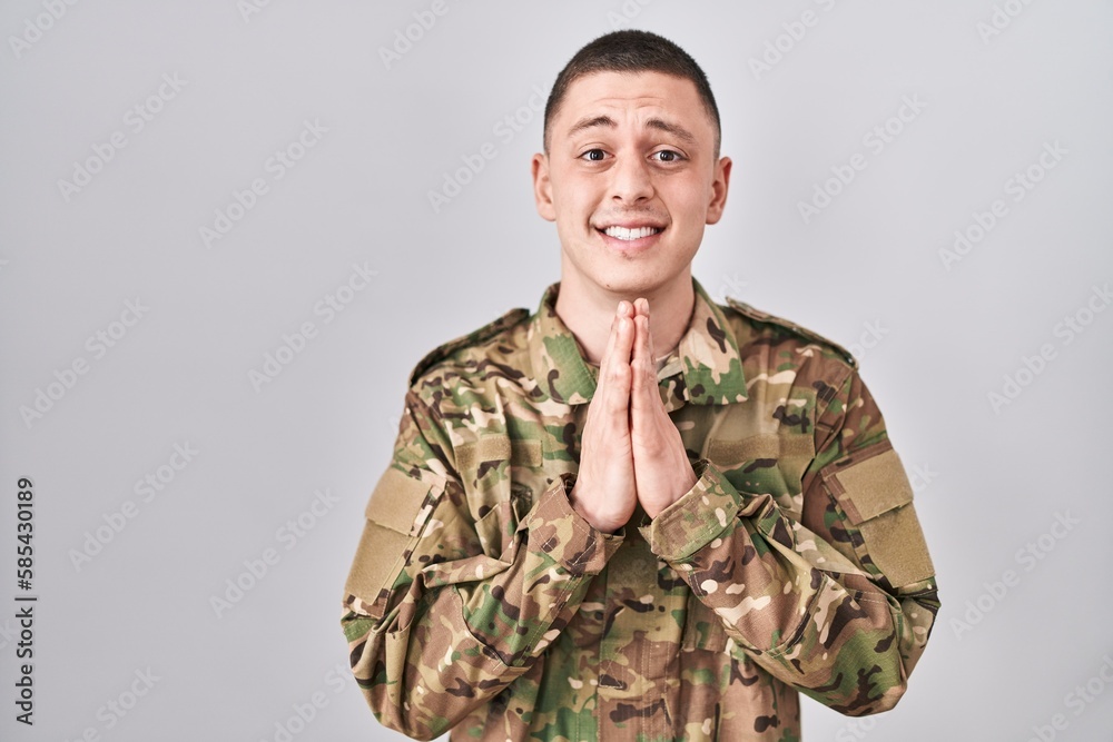 Young man wearing camouflage army uniform praying with hands together asking for forgiveness smiling confident.