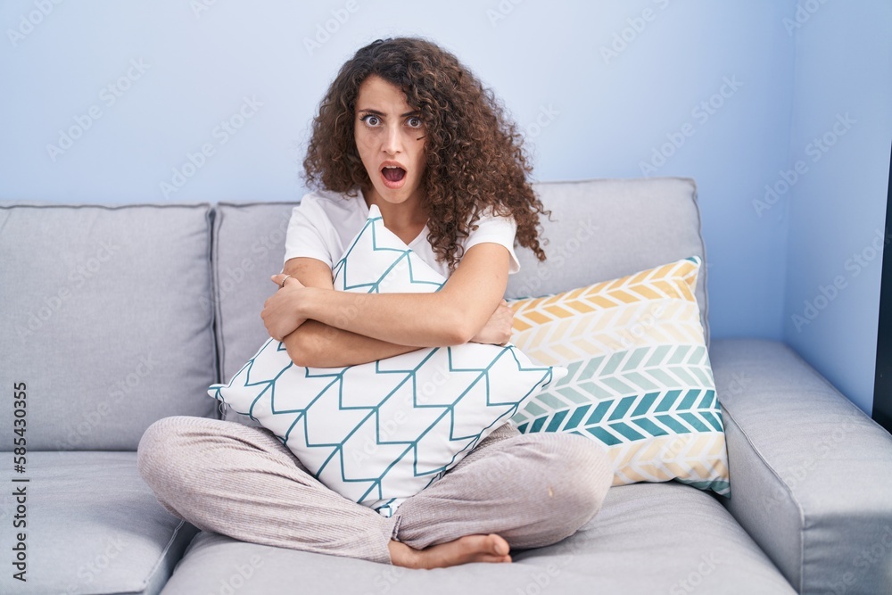 Hispanic woman with curly hair sitting on the sofa at home in shock face, looking skeptical and sarcastic, surprised with open mouth Hispanic woman with curly hair sitting on the sofa at home in shock face, looking skeptical and sarcastic, surprised with open mouth