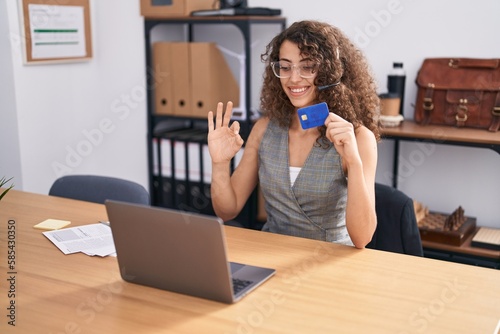 Hispanic woman with curly hair wearing call center agent headset holding credit card doing ok sign with fingers, smiling friendly gesturing excellent symbol