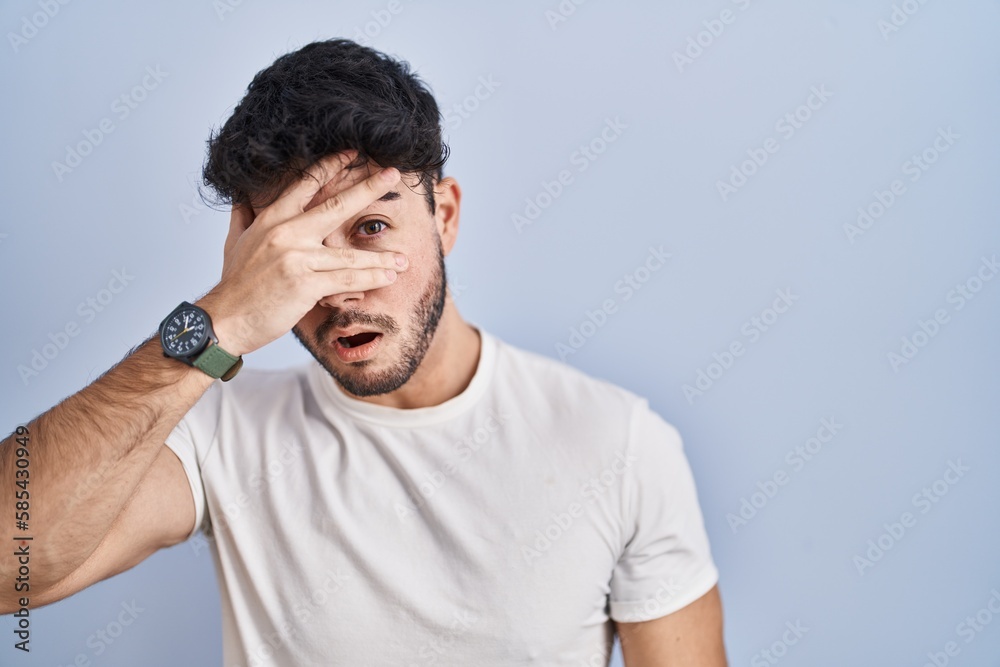 Hispanic man with beard standing over white background peeking in shock covering face and eyes with hand, looking through fingers with embarrassed expression. Hispanic man with beard standing over white background peeking in shock covering face and eyes with hand, looking through fingers with embarrassed expression.