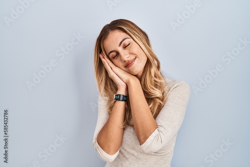 Young blonde woman standing over isolated background sleeping tired dreaming and posing with hands together while smiling with closed eyes.
