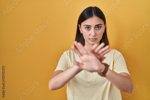 Hispanic girl wearing casual t shirt over yellow background rejection expression crossing arms and palms doing negative sign, angry face
