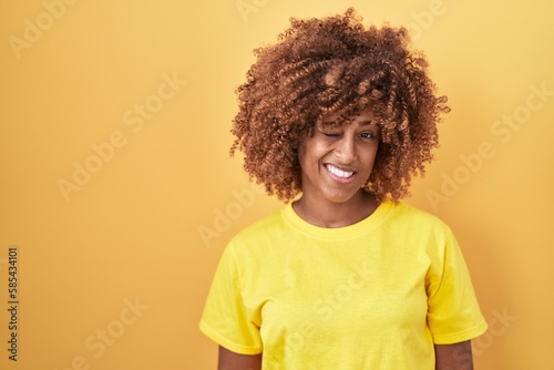 Young hispanic woman with curly hair standing over yellow background winking looking at the camera with sexy expression, cheerful and happy face.