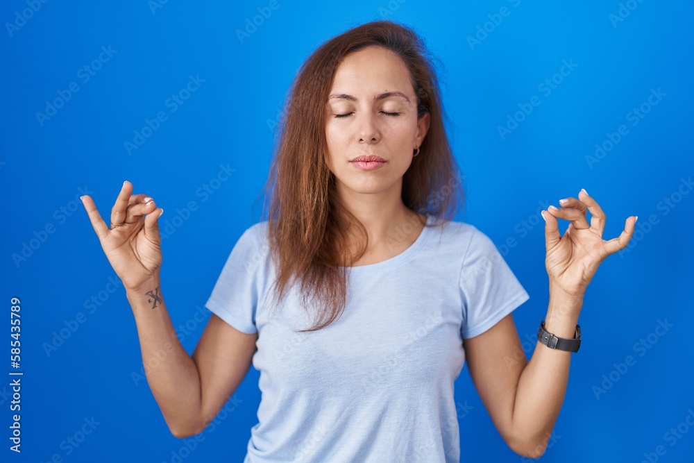 Brunette woman standing over blue background relaxed and smiling with eyes closed doing meditation gesture with fingers. yoga concept. Brunette woman standing over blue background relaxed and smiling with eyes closed doing meditation gesture with fingers. yoga concept.