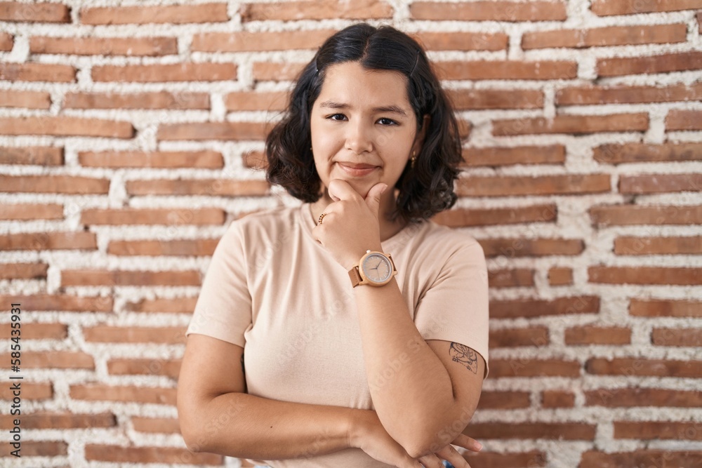 Young hispanic woman standing over bricks wall looking confident at the camera smiling with crossed arms and hand raised on chin. thinking positive. Young hispanic woman standing over bricks wall looking confident at the camera smiling with crossed arms and hand raised on chin. thinking positive.