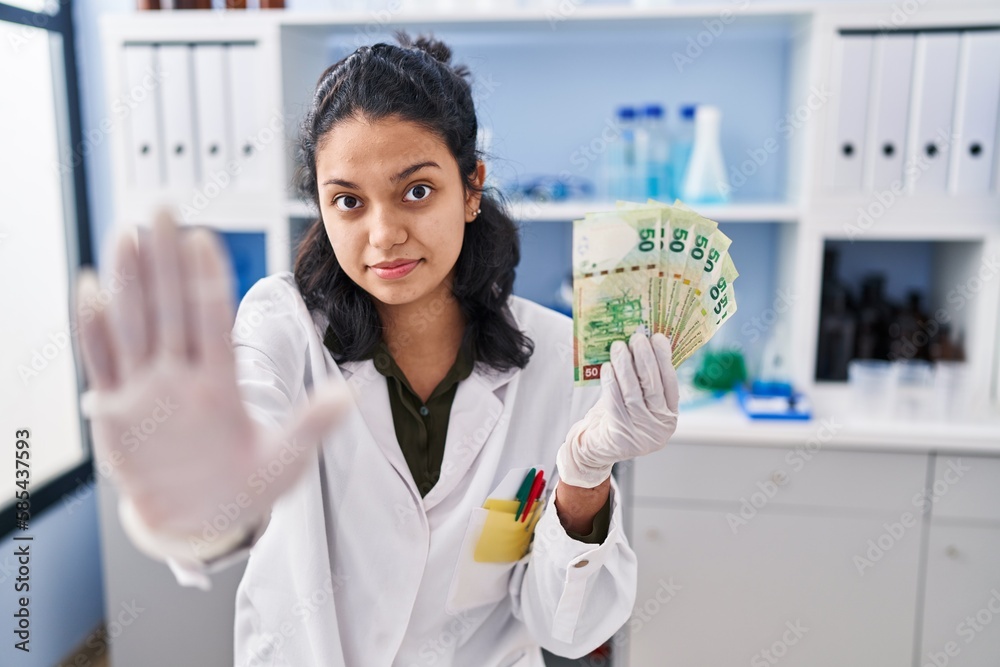 Hispanic woman with dark hair working at scientist laboratory holding money with open hand doing stop sign with serious and confident expression, defense gesture